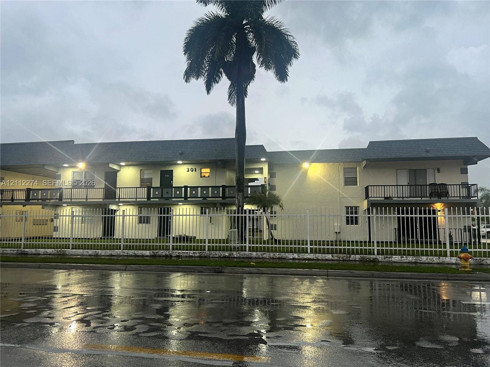 301 NW 177th Street, #141, Miami Gardens, FL 33169 Multi-level exterior featuring a light-colored stucco facade, shingled roofing, second-story railed balconies, and perimeter metal fencing