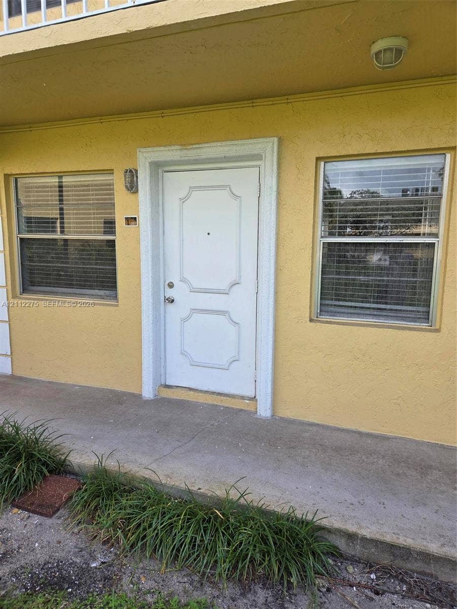 Exterior entryway featuring a white paneled door with decorative molding, two windows with horizontal blinds, a concrete porch, and an overhead flush-mount light fixture