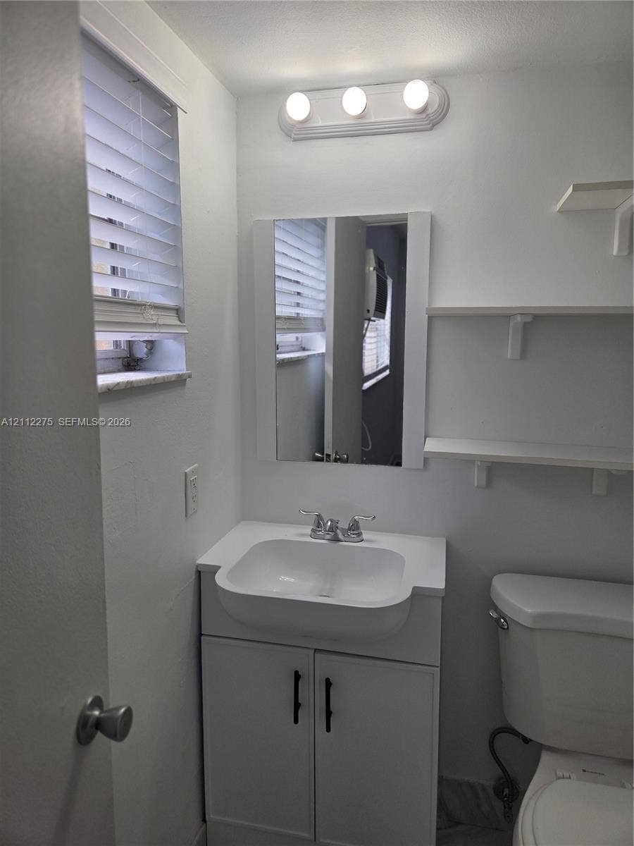 White vanity with a rectangular sink, chrome dual-handle faucet, frameless mirror, three-light fixture, and wall-mounted shelving