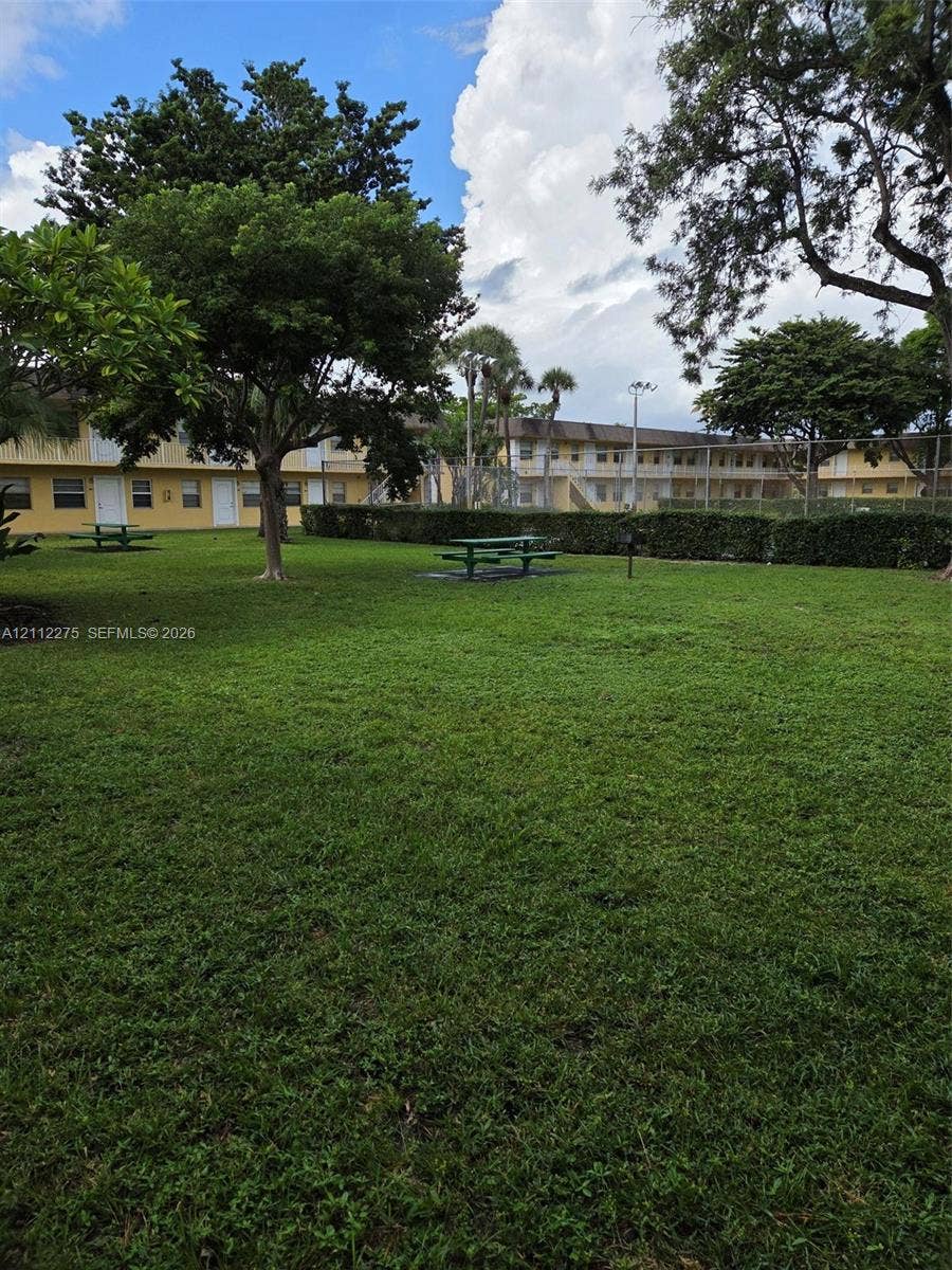 Expansive grassy common area featuring mature shade trees, perimeter hedging, and fixed metal picnic tables