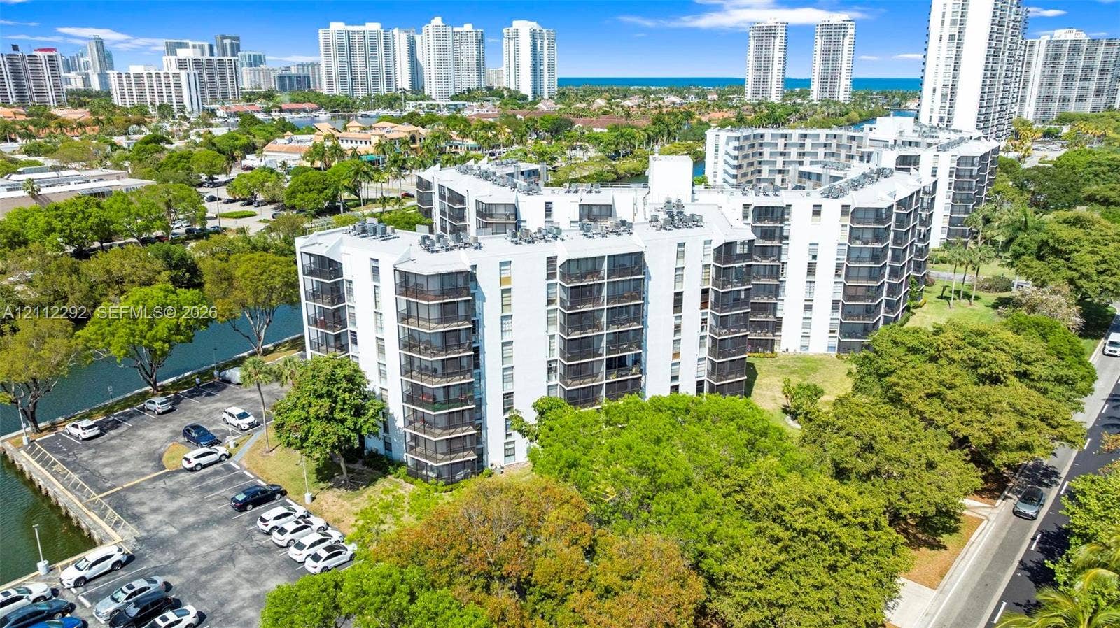 Multi-story building featuring screened balconies, water-adjacent hardscaping, paved parking, and landscaped grounds