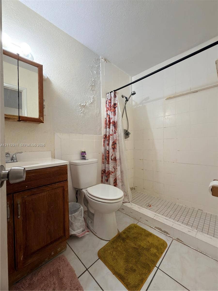 390 NW 49th Avenue, Plantation, FL 33317 Bathroom featuring a wood-finish vanity with a single-basin sink, wall-mounted medicine cabinet, white tile wall surround, tile shower flooring, and ceramic floor tile
