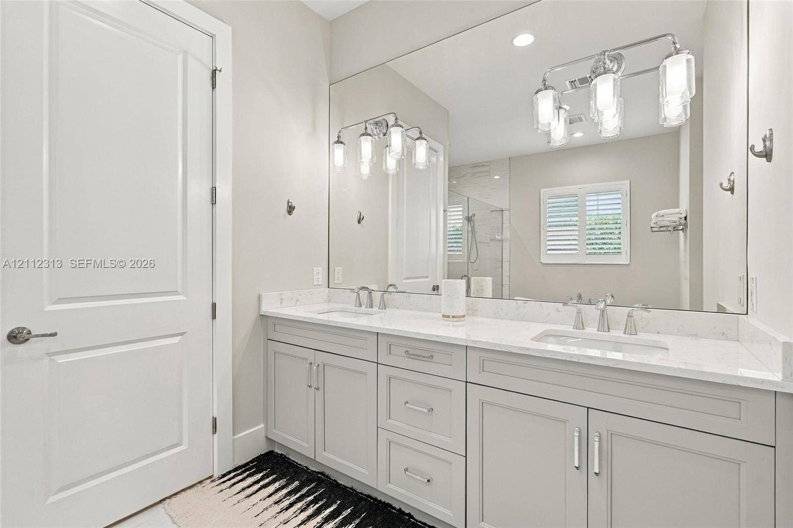 Double vanity featuring light-toned cabinetry, stone-finish countertops, undermount sinks, polished metal fixtures, and a wall-to-wall mirror with multi-bulb integrated lighting