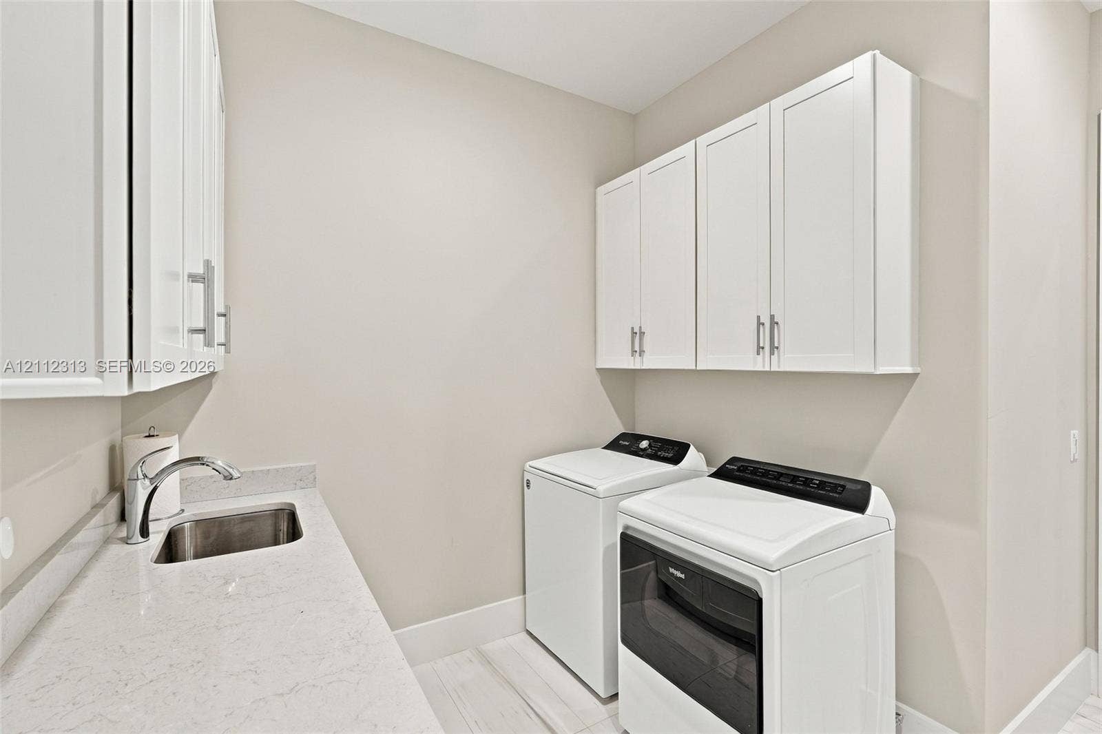 Laundry area featuring white shaker-style wall cabinets, a light-toned countertop, an integrated stainless steel sink with a chrome faucet, and wood-finish flooring