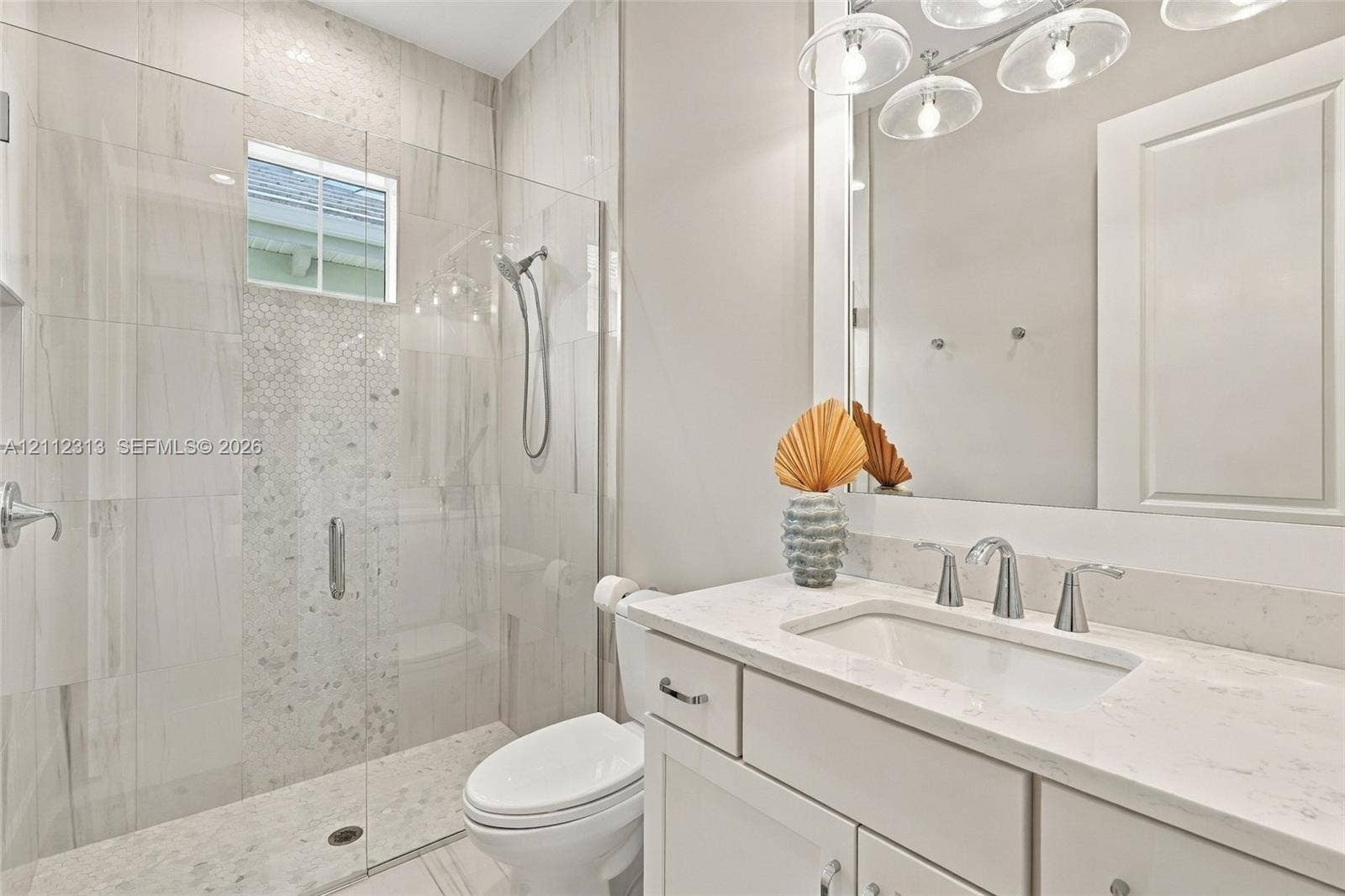 Bathroom featuring a vanity with a white countertop and undermount sink, a walk-in shower with a glass enclosure, floor-to-ceiling tilework, and a multi-bulb light fixture