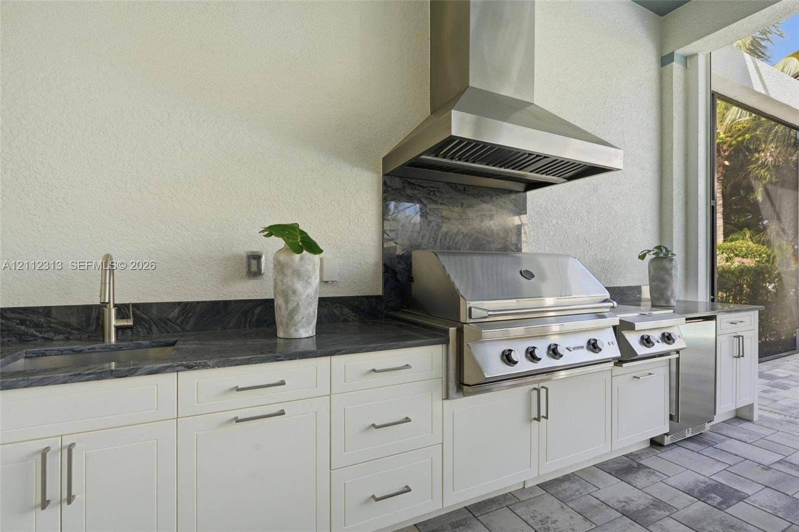 Outdoor kitchen featuring white shaker-style cabinetry, dark natural stone countertops and backsplash, stainless steel built-in grill, commercial-grade ventilation hood, and under-mount sink with modern faucet