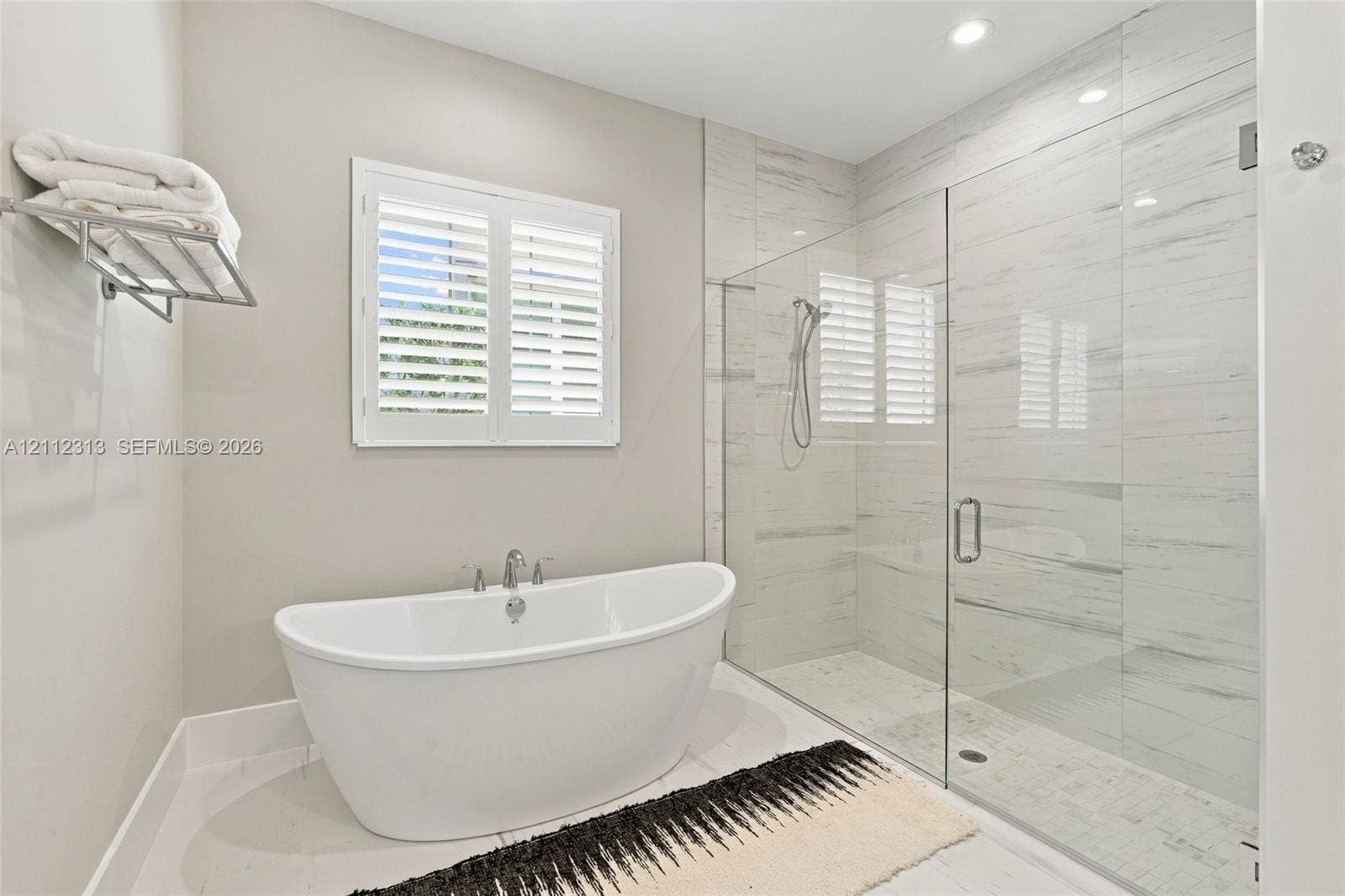 Freestanding soaking tub with chrome fixtures, glass-enclosed walk-in shower with floor-to-ceiling tile, and window shutters
