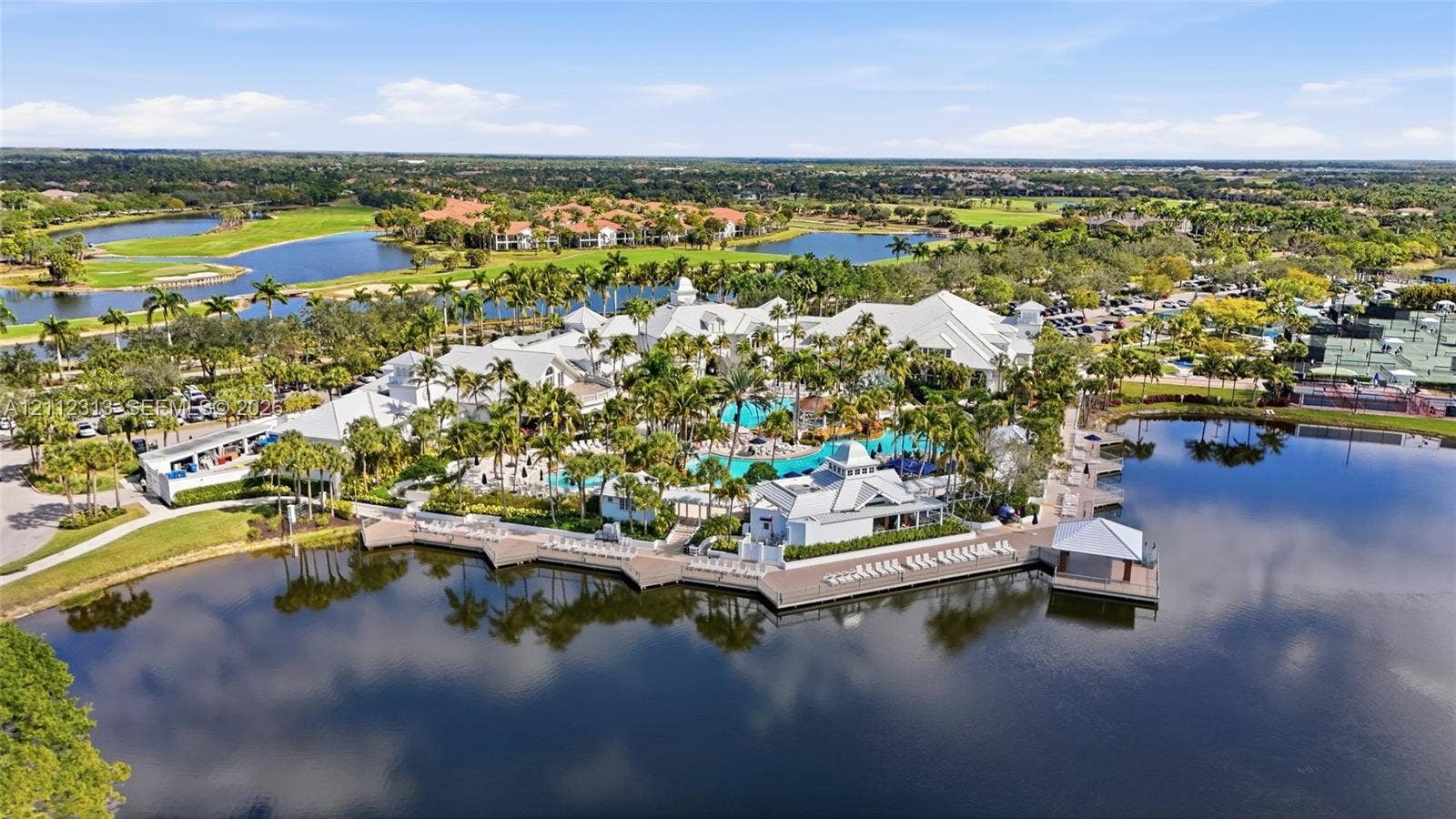 Aerial view of a coastal-inspired complex featuring a resort-style swimming pool, expansive waterfront sun deck, multi-tiered white roofing, and manicured tropical landscaping