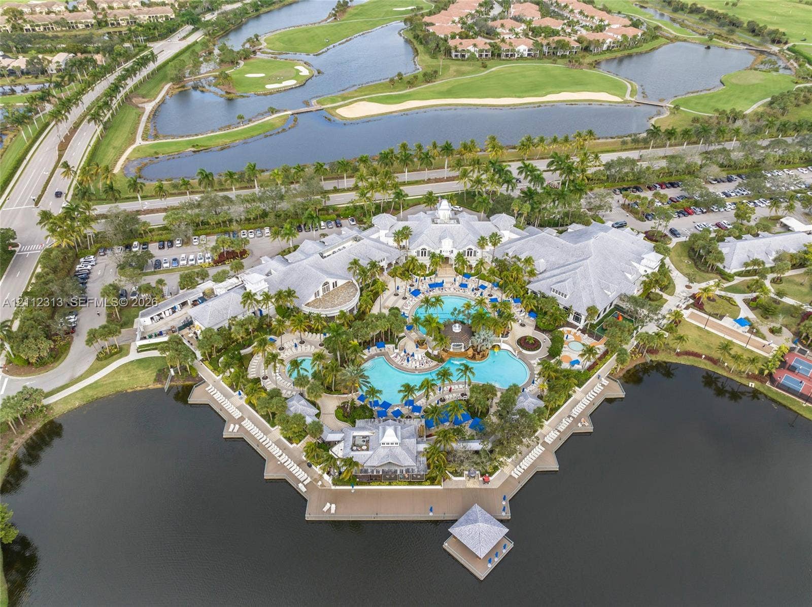 Aerial perspective of a multi-structure facility featuring a complex of resort-style swimming pools, expansive paved sun decks, a waterfront gazebo, and mature landscaping situated along a large lake
