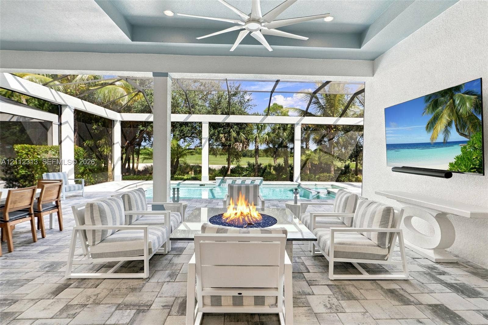Covered outdoor living space featuring a recessed tray ceiling, multi-blade ceiling fan, paver flooring, built-in wall-mounted television, and a screen-enclosed pool area