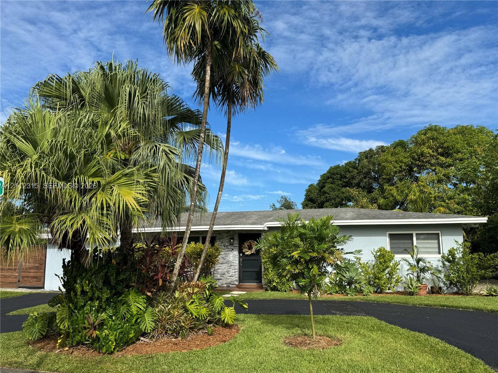Single-story exterior featuring light-toned horizontal siding, a stone veneer facade, an asphalt driveway, and mature palm trees