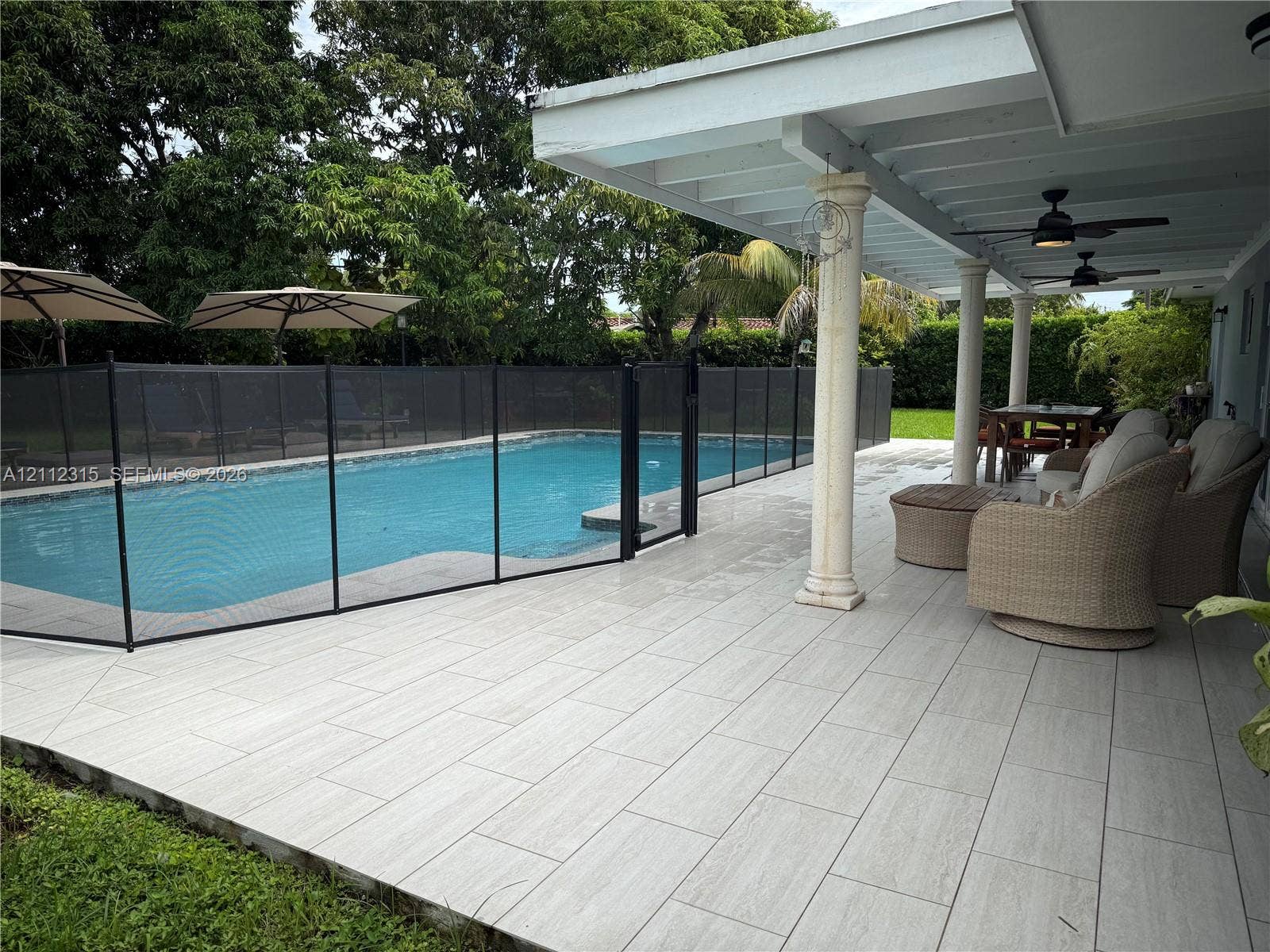 Covered patio with large-format tile flooring and decorative columns, featuring a swimming pool, safety fencing, and mature perimeter landscaping