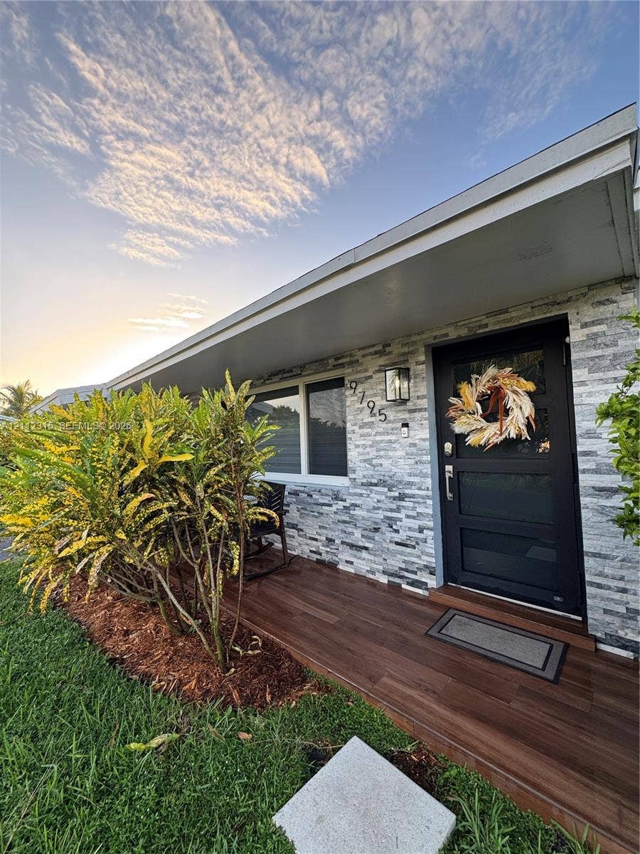 Exterior facade featuring stacked stone siding, wood-finish porch flooring, a modern multi-panel entry door, and wall-mounted exterior lighting