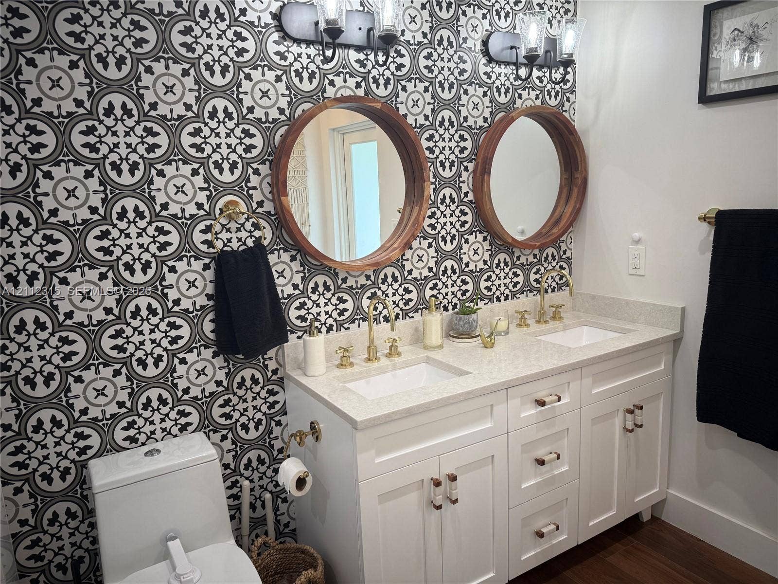 Dual-sink vanity with white cabinetry, quartz countertop, round wood-frame mirrors, geometric patterned wall tile, and brass-tone fixtures