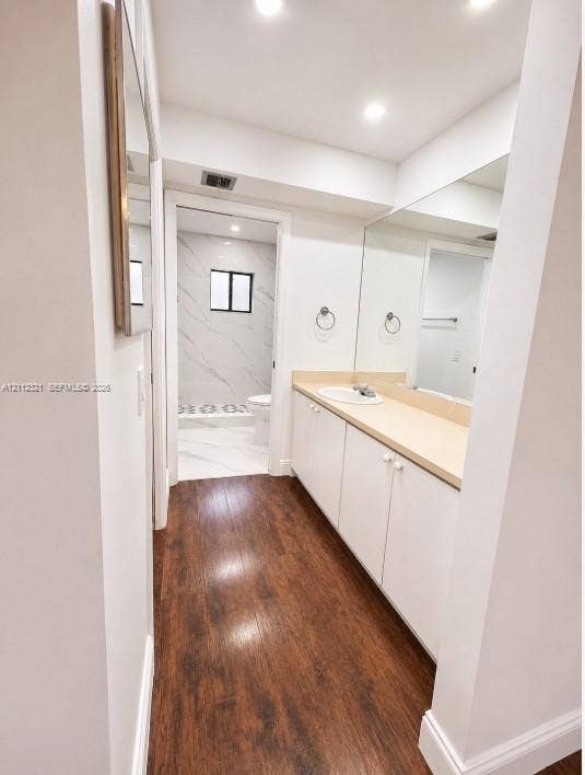 Bathroom featuring wood-finish flooring, white cabinetry, a solid-surface countertop with an integrated sink, full-width mirror, and recessed lighting