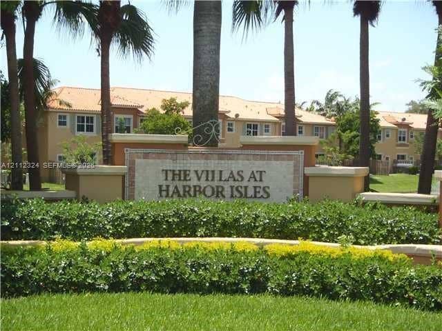 Community monument sign featuring stucco and stone masonry construction, accented by manicured hedges and mature palm trees