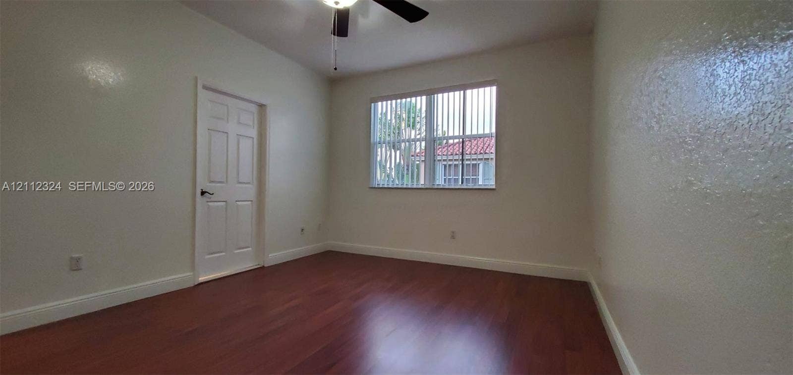 Interior room featuring wood-finish flooring, a multi-pane window with vertical blinds, a six-panel interior door, and a ceiling-mounted light fixture with fan