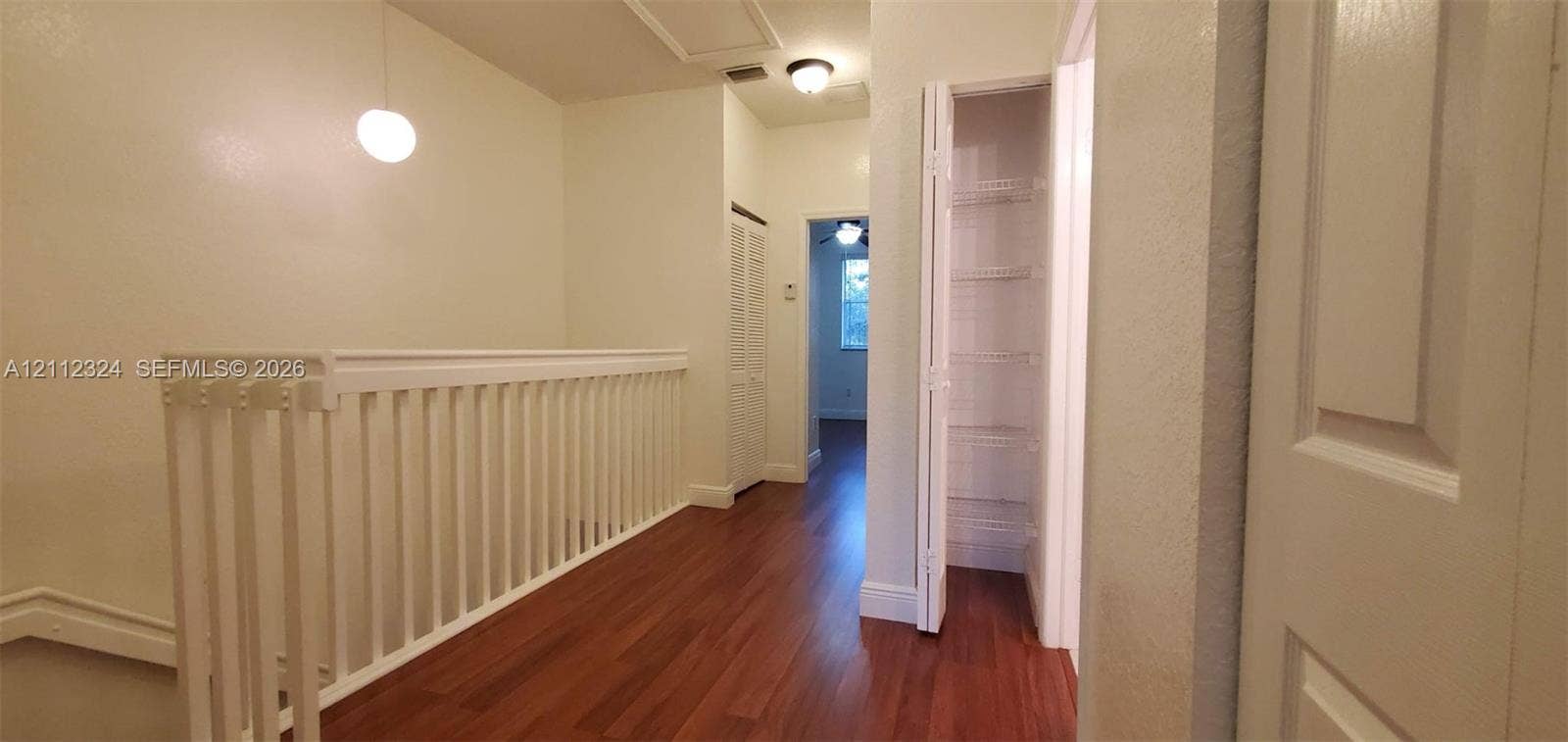 Hallway featuring wood-finish flooring, white spindle railing, built-in louvered-door closet, and modern pendant lighting