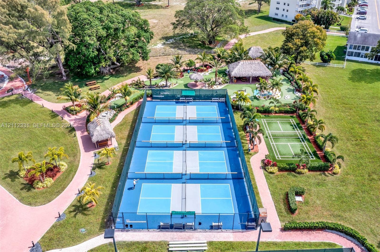4025 Upminster K, #4025, Deerfield Beach, FL 33442 Recreational complex featuring four pickleball courts, a grass tennis court, multiple thatch-roofed pavilions, and landscaped walking paths