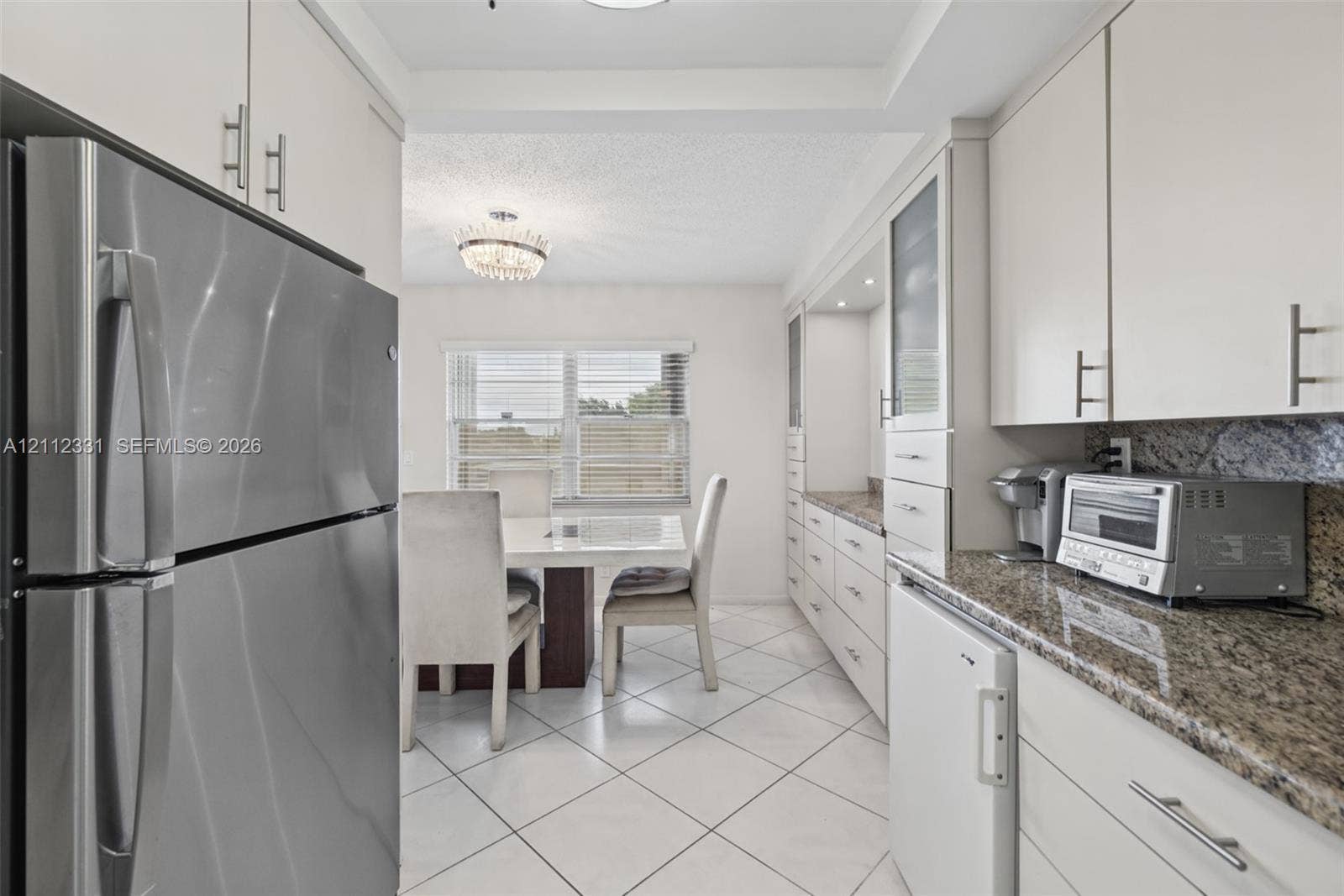 4025 Upminster K, #4025, Deerfield Beach, FL 33442 Kitchen featuring white cabinetry with brushed metal hardware, speckled stone countertops, stainless steel refrigerator, white ceramic tile flooring, and a crystal-accented chandelier