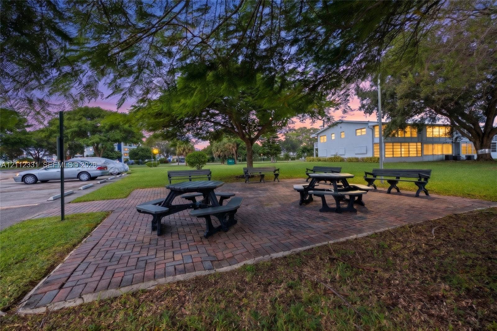 4025 Upminster K, #4025, Deerfield Beach, FL 33442 Outdoor common area featuring a herringbone-patterned brick paver patio, permanent benches, and mature shade trees set within a landscaped lawn