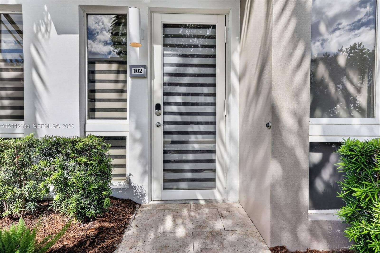 10305 NW 63rd Terrace, #102, Doral, FL 33178 Exterior entry featuring a white door with horizontal glass panes, stone-finish paving, modern cylindrical wall-mounted lighting, and manicured shrubbery