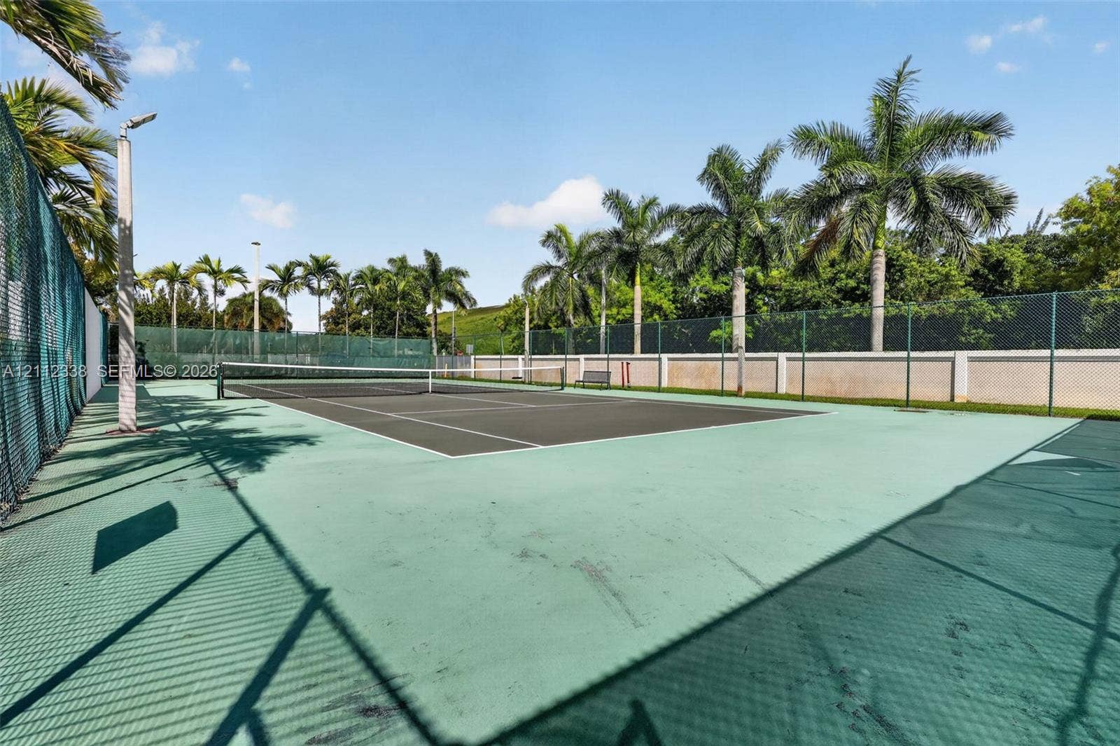 10305 NW 63rd Terrace, #102, Doral, FL 33178 Tennis court featuring a paved playing surface, perimeter chain-link fencing, high-mast lighting, and mature palm trees