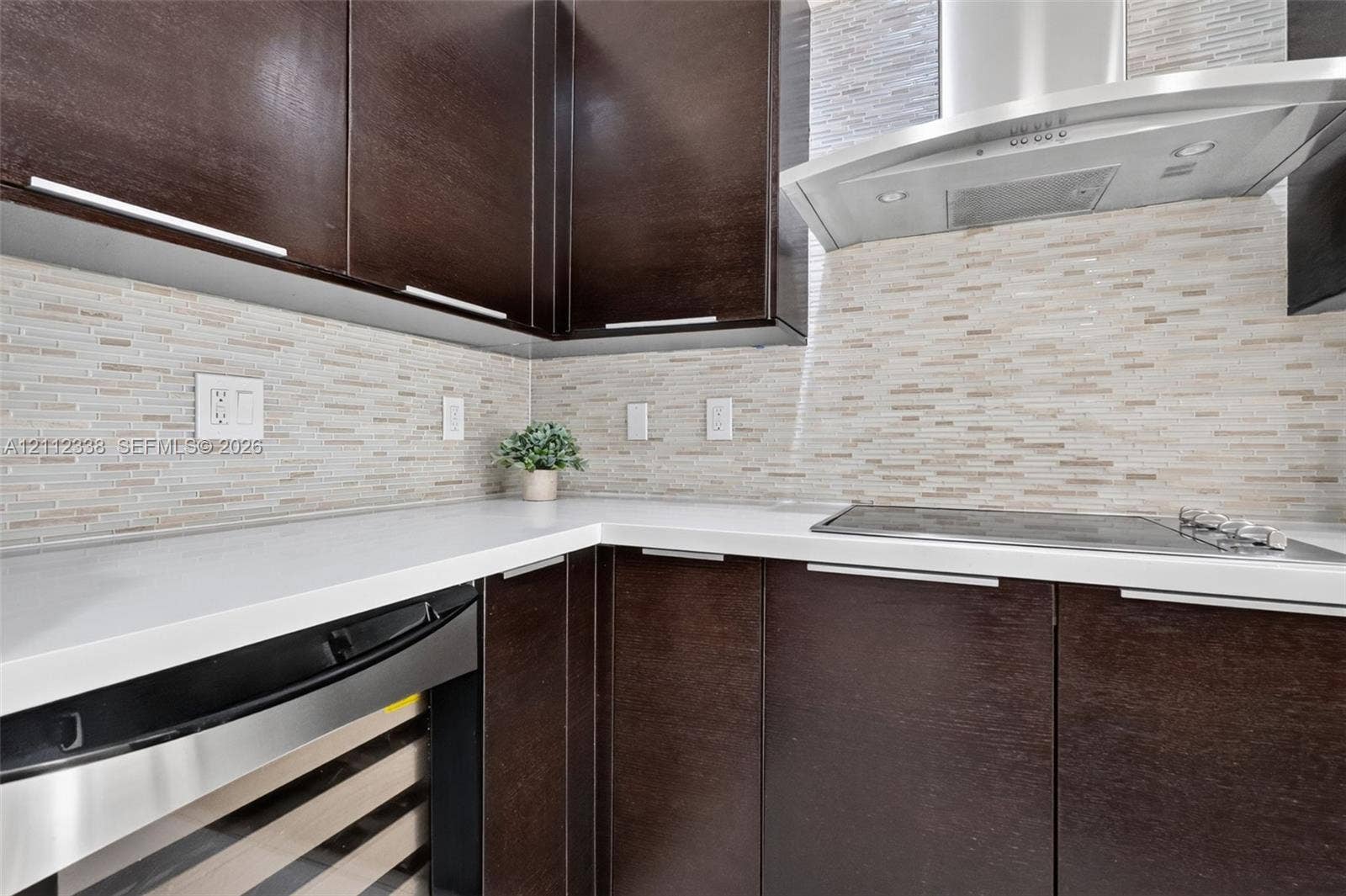 10305 NW 63rd Terrace, #102, Doral, FL 33178 Dark wood-finish cabinetry featuring sleek hardware, white solid-surface countertops, horizontal linear tile backsplash, stainless steel range hood, and integrated under-counter beverage chiller