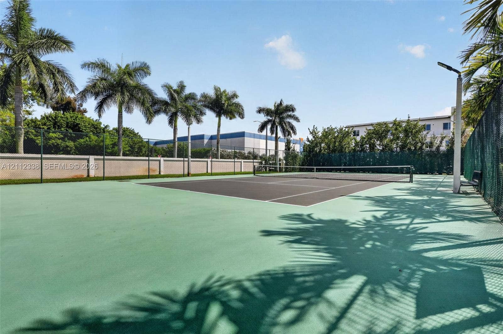 10305 NW 63rd Terrace, #102, Doral, FL 33178 Tennis court with a high-contrast playing surface, enclosed by chain-link fencing, featuring overhead lighting and mature palm tree landscaping