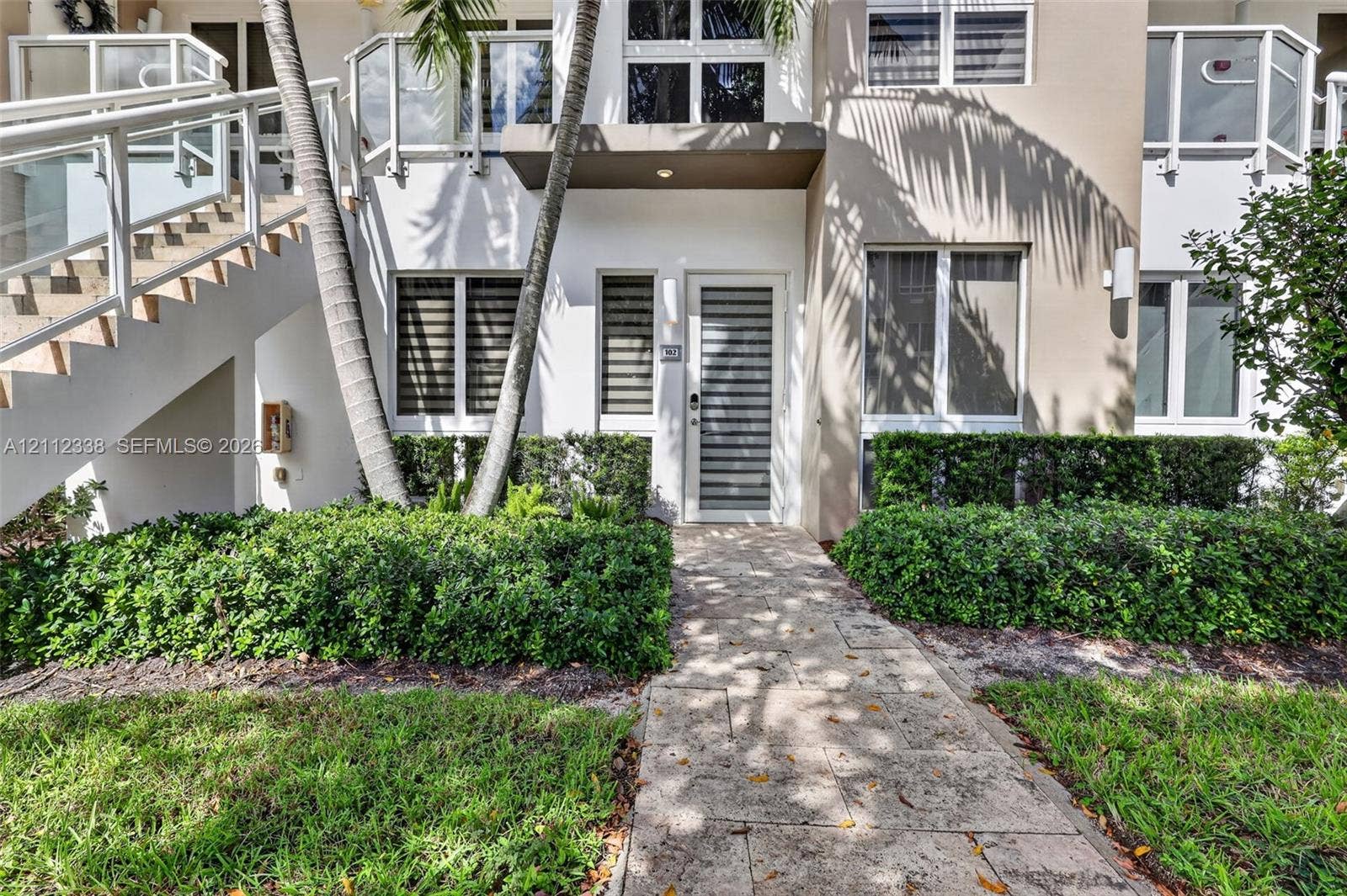 10305 NW 63rd Terrace, #102, Doral, FL 33178 Exterior featuring a stone paver walkway, multi-pane windows with horizontal blinds, glass-panel stair railing, and a private entrance with a horizontal-slat door