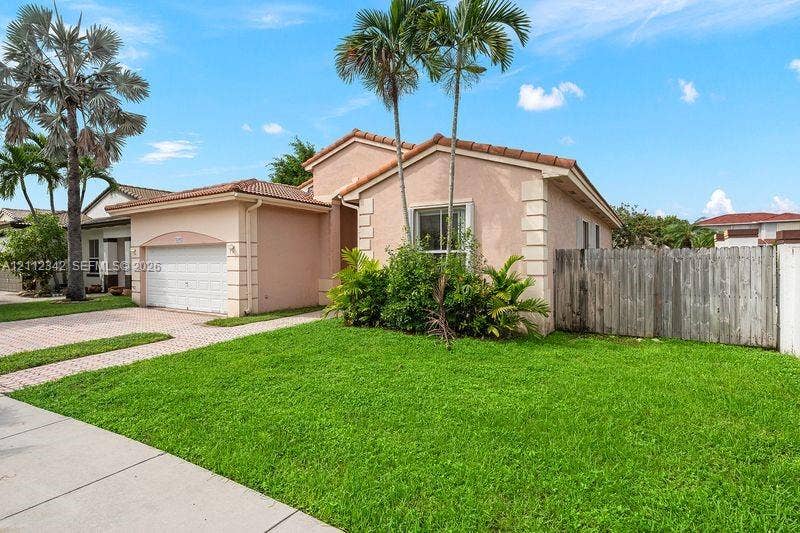 Single-story exterior featuring a stucco facade, tile roof, paver driveway, attached two-car garage, and wood-panel perimeter fencing