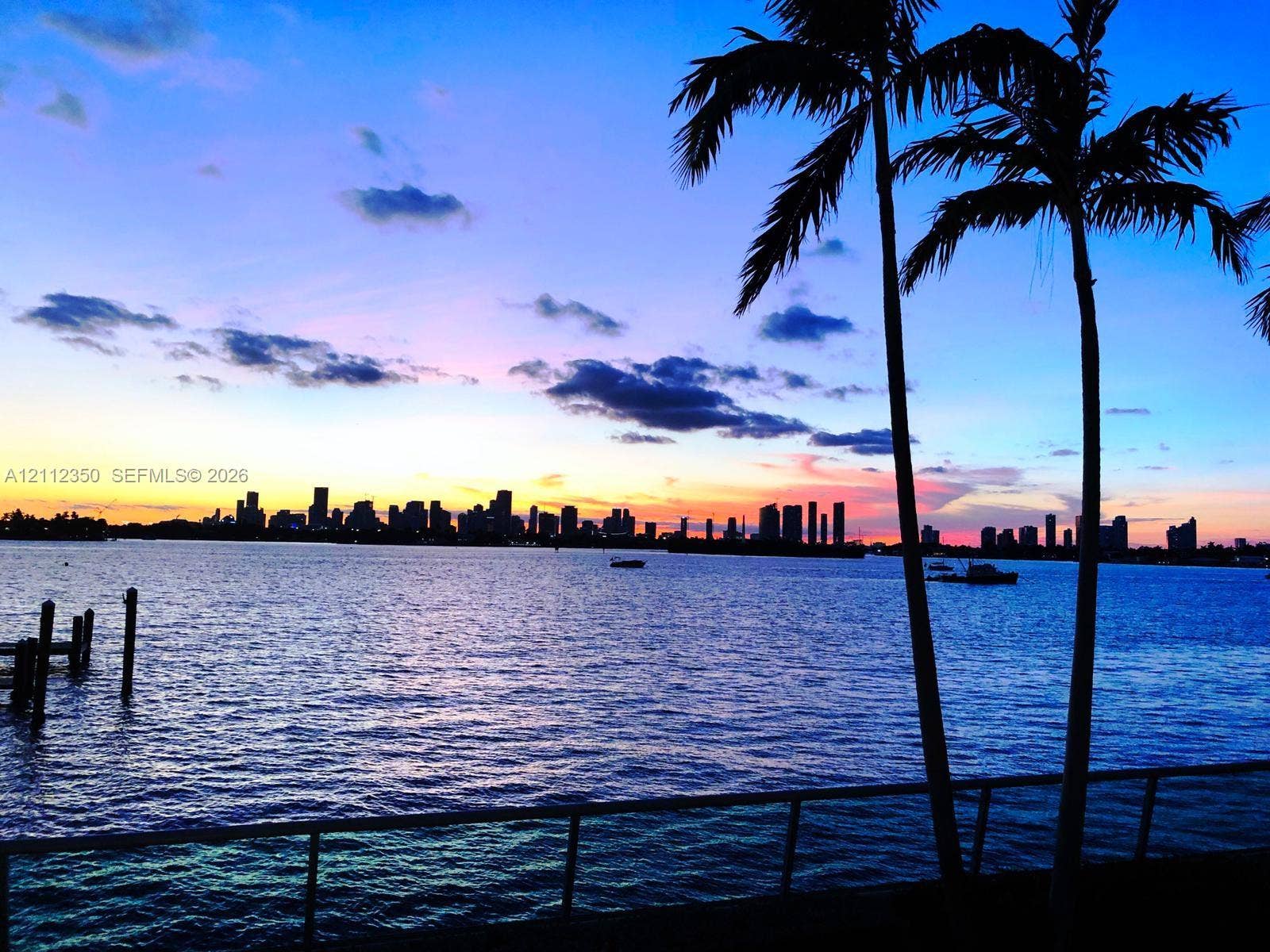 Waterfront property featuring a seawall, metal railing, and mature palm trees with a city skyline backdrop