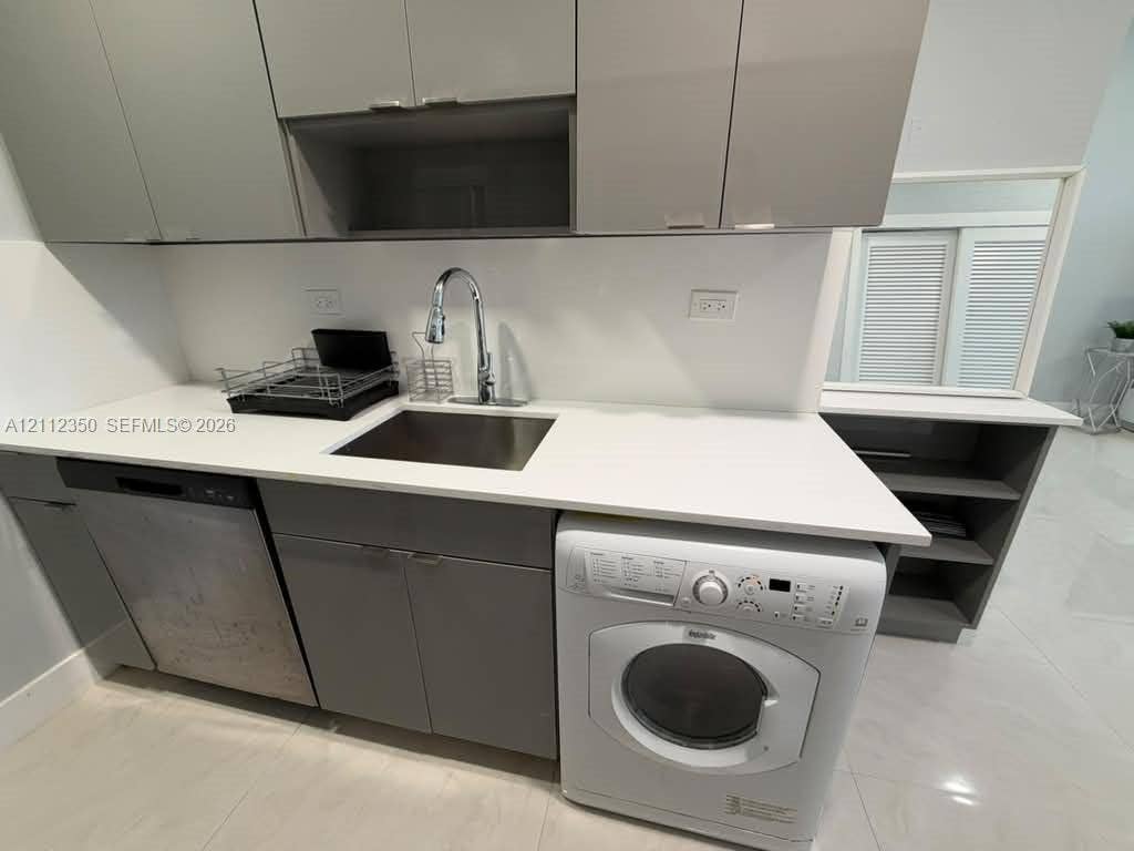 Contemporary cabinetry with grey finish, solid surface countertop, stainless steel sink with high-arc faucet, built-in dishwasher, and front-loading laundry unit