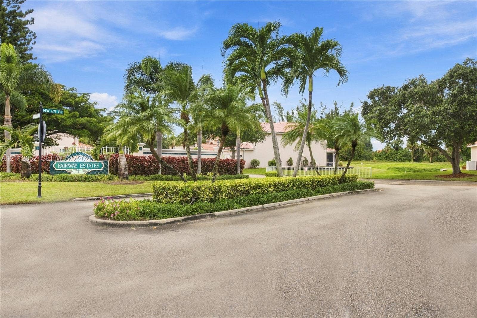 2880 NW 75th Avenue, Margate, FL 33063 Entryway featuring mature palm trees, manicured hedges, paved roadways, and ornamental shrubbery