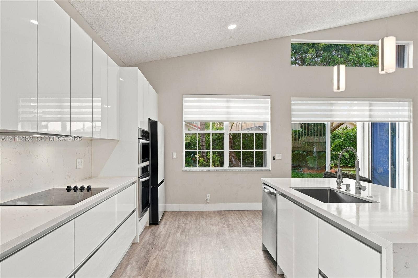 2880 NW 75th Avenue, Margate, FL 33063 Modern kitchen featuring high-gloss white cabinetry, quartz countertops, a central island with a stainless steel sink, wood-finish flooring, and vaulted ceilings with clerestory windows