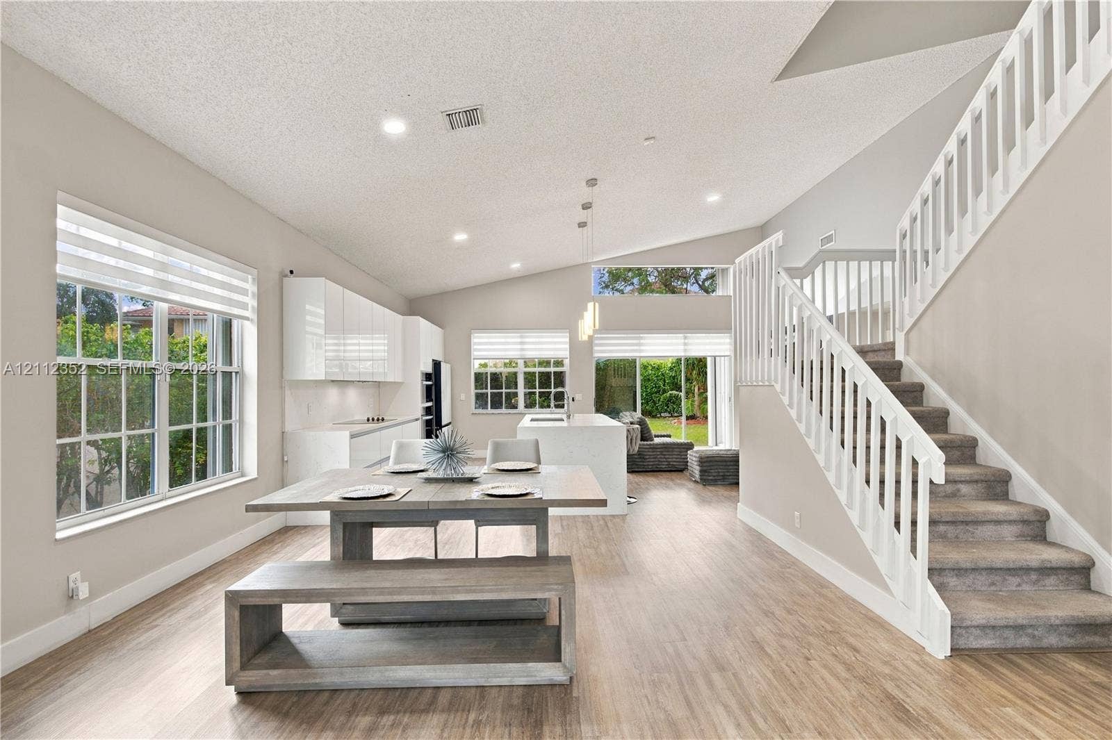 2880 NW 75th Avenue, Margate, FL 33063 Open-concept interior featuring wood-finish flooring, vaulted ceilings, high-gloss white cabinetry, a center kitchen island with waterfall-edge countertop, and a staircase with white balusters