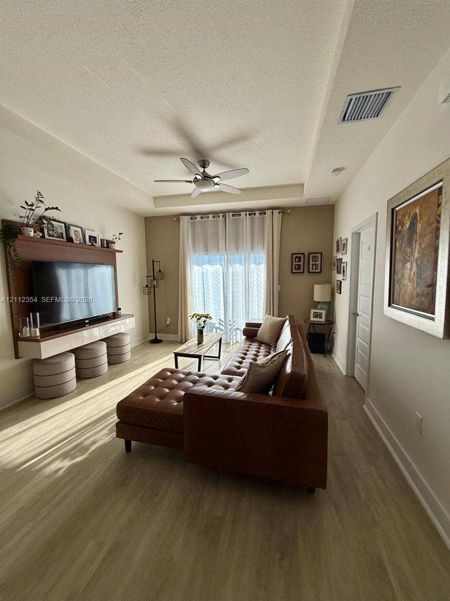 Tray ceiling with a central multi-blade ceiling fan, wood-finish flooring, and a wall-mounted built-in media console with integrated shelving