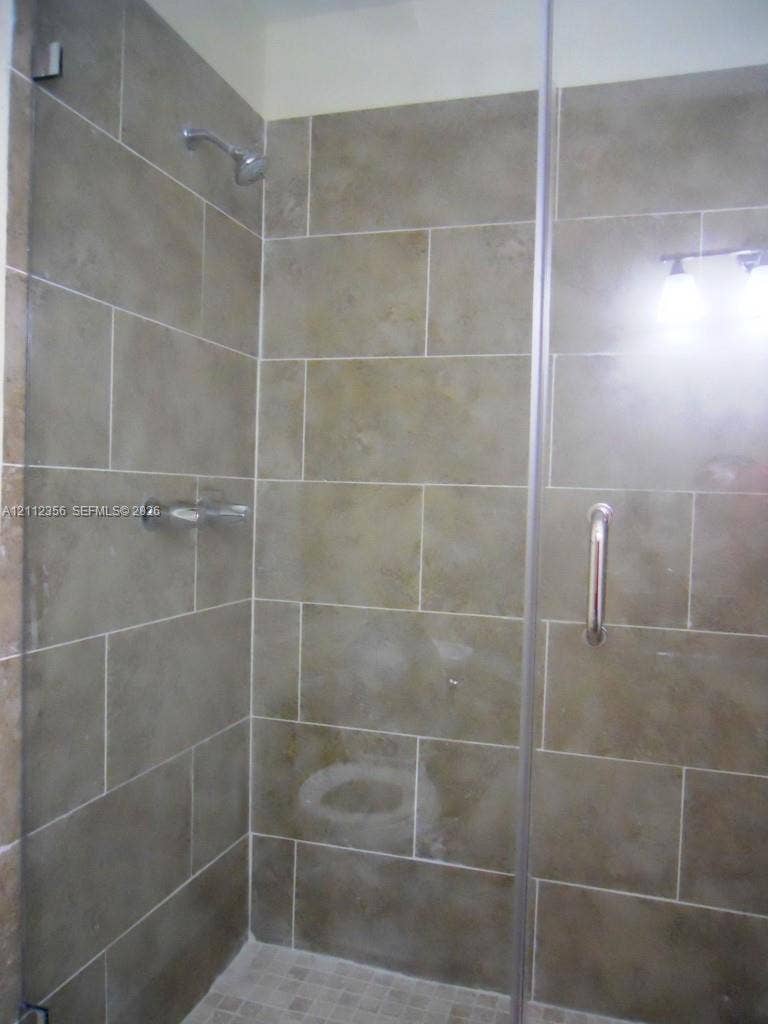 Walk-in shower featuring large-format neutral tile wall surround, glass enclosure with brushed metal hardware, wall-mounted showerhead, and mosaic tile flooring