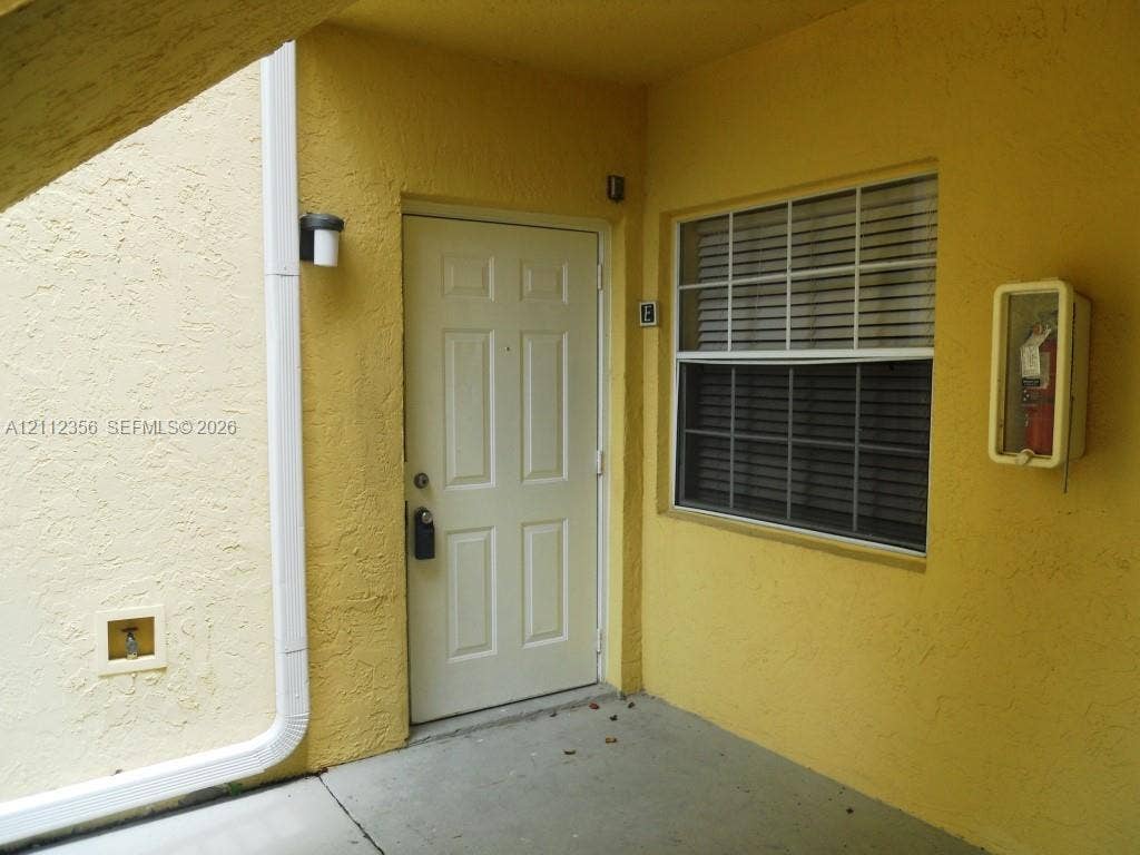 Entryway featuring a solid-panel door, grid-pattern window, stucco exterior finish, wall-mounted light fixture, and recessed utility access