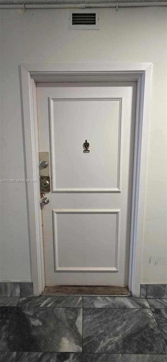 White paneled door featuring a decorative metal door knocker, dual deadbolt locks, and a polished metal door handle, set against stone-finish flooring
