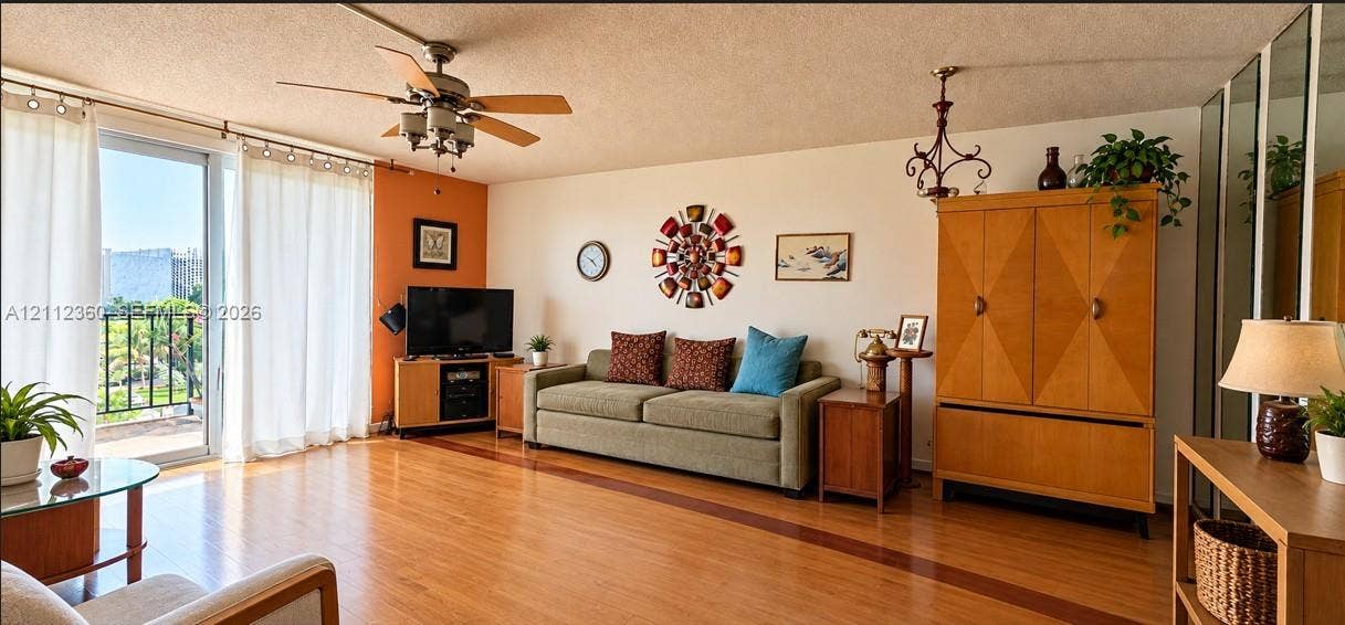 Living area featuring wood-finish flooring, a ceiling fan with integrated lighting, sliding glass door access to a balcony, and mirrored closet doors