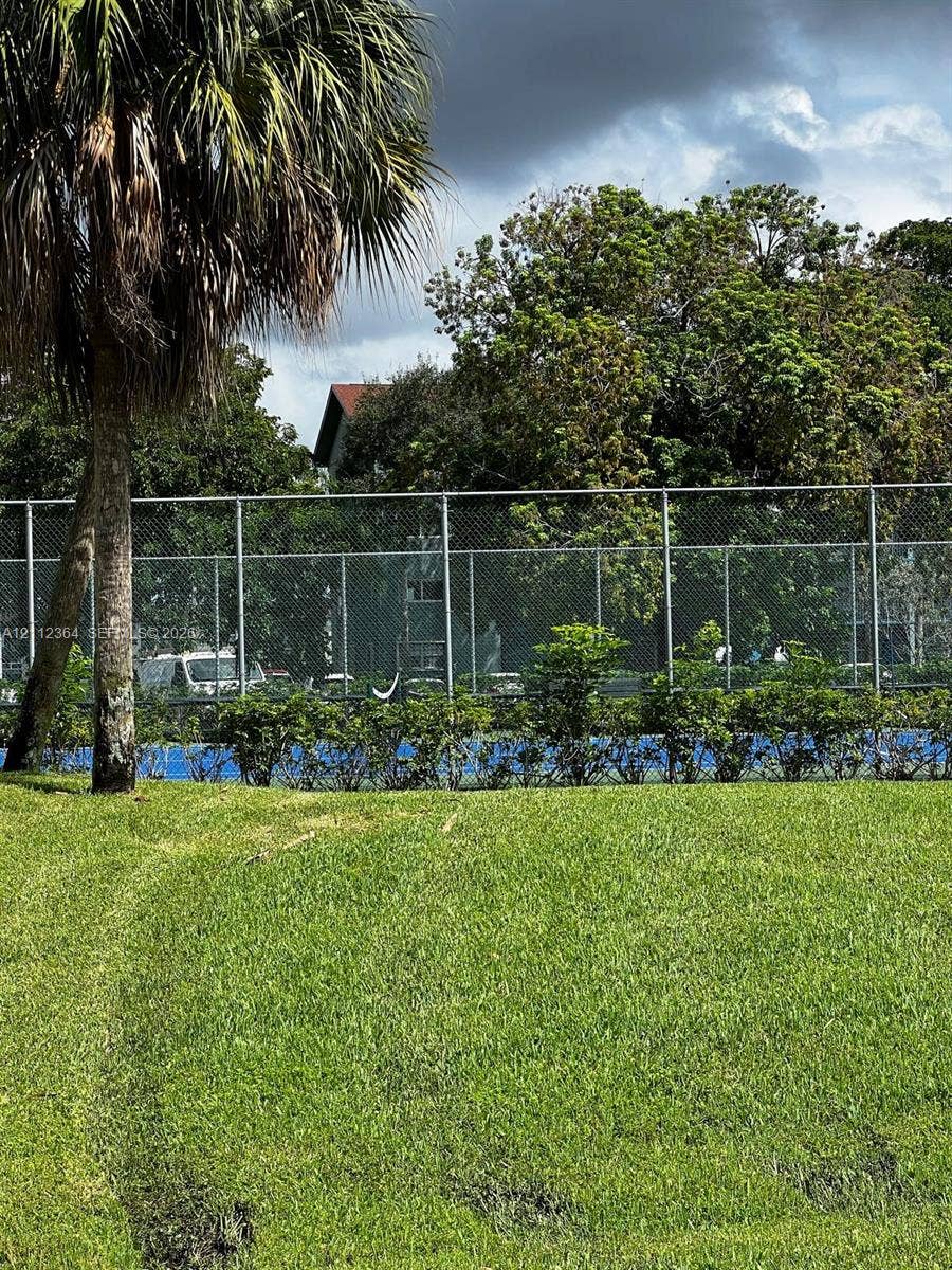 Recreational tennis court featuring a perimeter chain-link fence, surrounded by mature trees and manicured lawn space