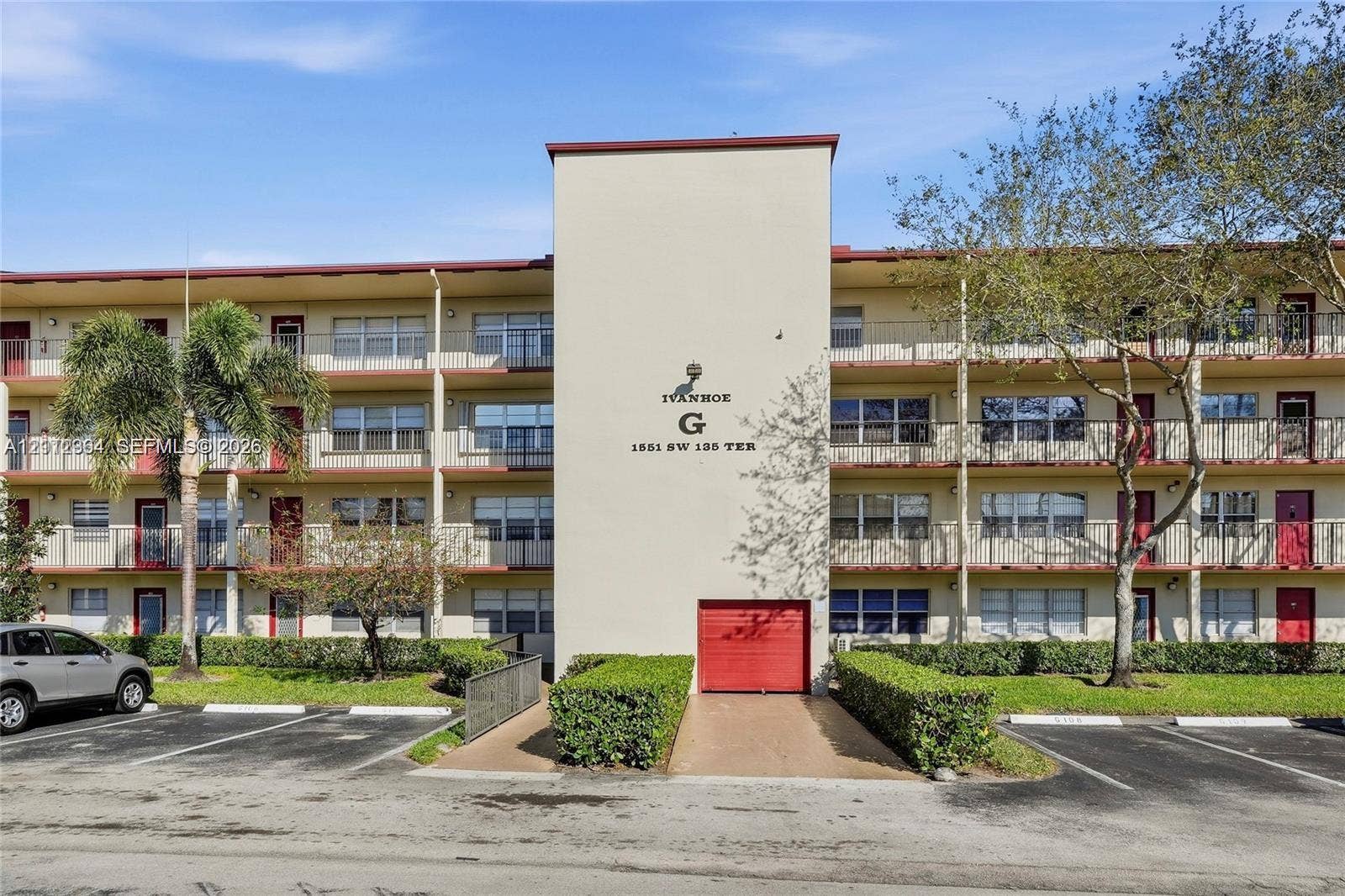 Multi-story residential building exterior featuring a central stucco facade, horizontal balconies with metal railings, red-accented doors, and asphalt parking area