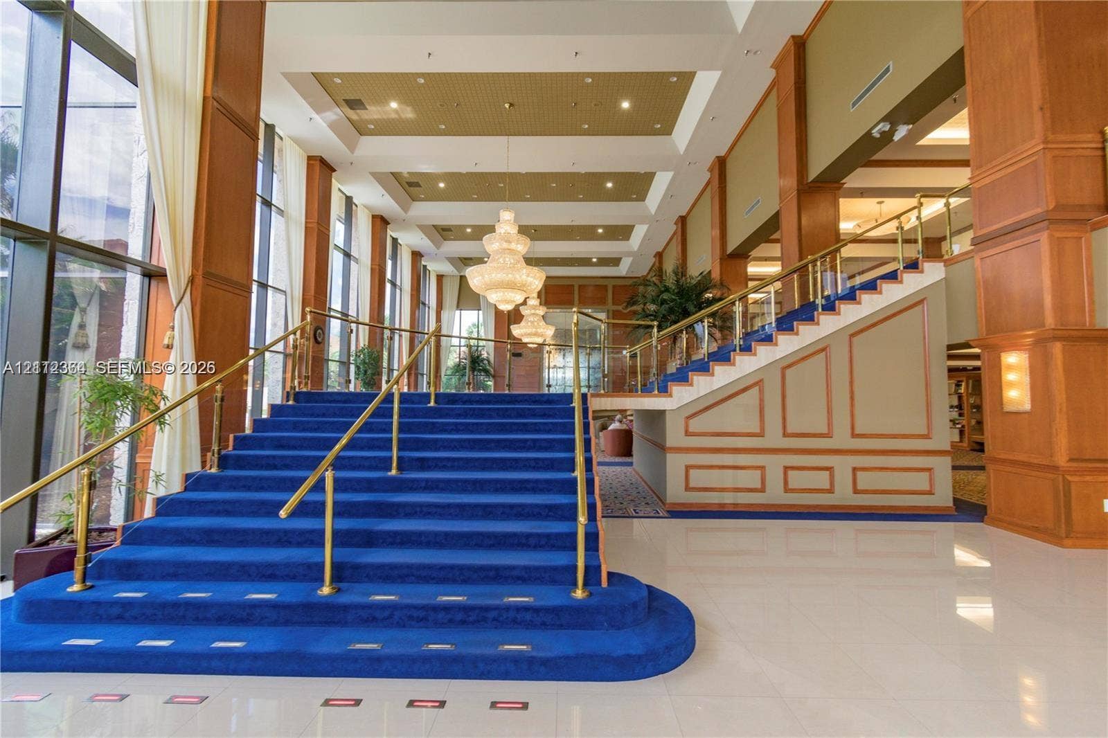 Grand entrance featuring a central staircase with blue carpeting and metal railings, high ceilings with recessed tray detailing, large floor-to-ceiling windows, tiered crystal chandeliers, and polished tile flooring