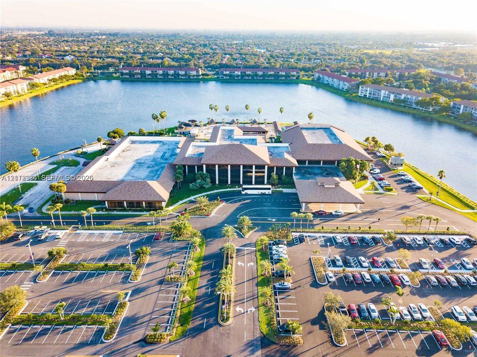 Aerial perspective of a large-scale commercial structure featuring a sprawling complex with multiple hipped roof sections, a dedicated covered arrival portico, an expansive paved parking facility, and surrounding water frontage