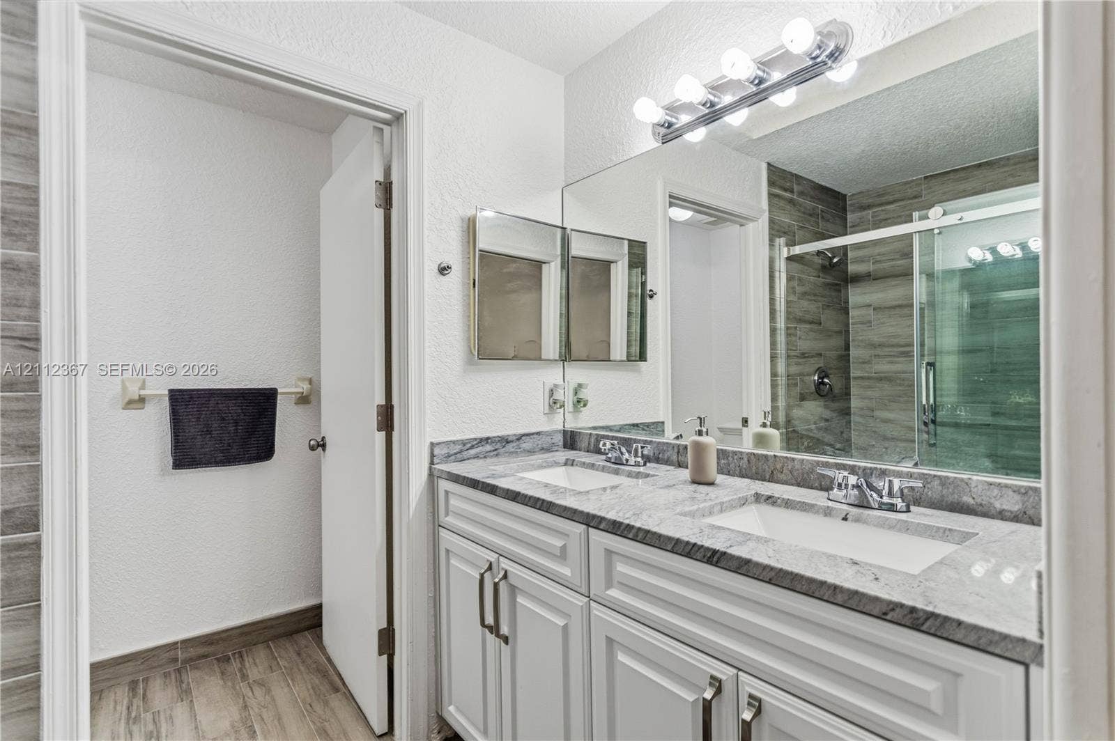 10803 Cypress Glen Drive, #10803, Coral Springs, FL 33071 Dual-sink vanity with stone-finish countertop, white shaker-style cabinetry, wood-finish flooring, full-width wall mirror, and shower enclosure with horizontal tile surround