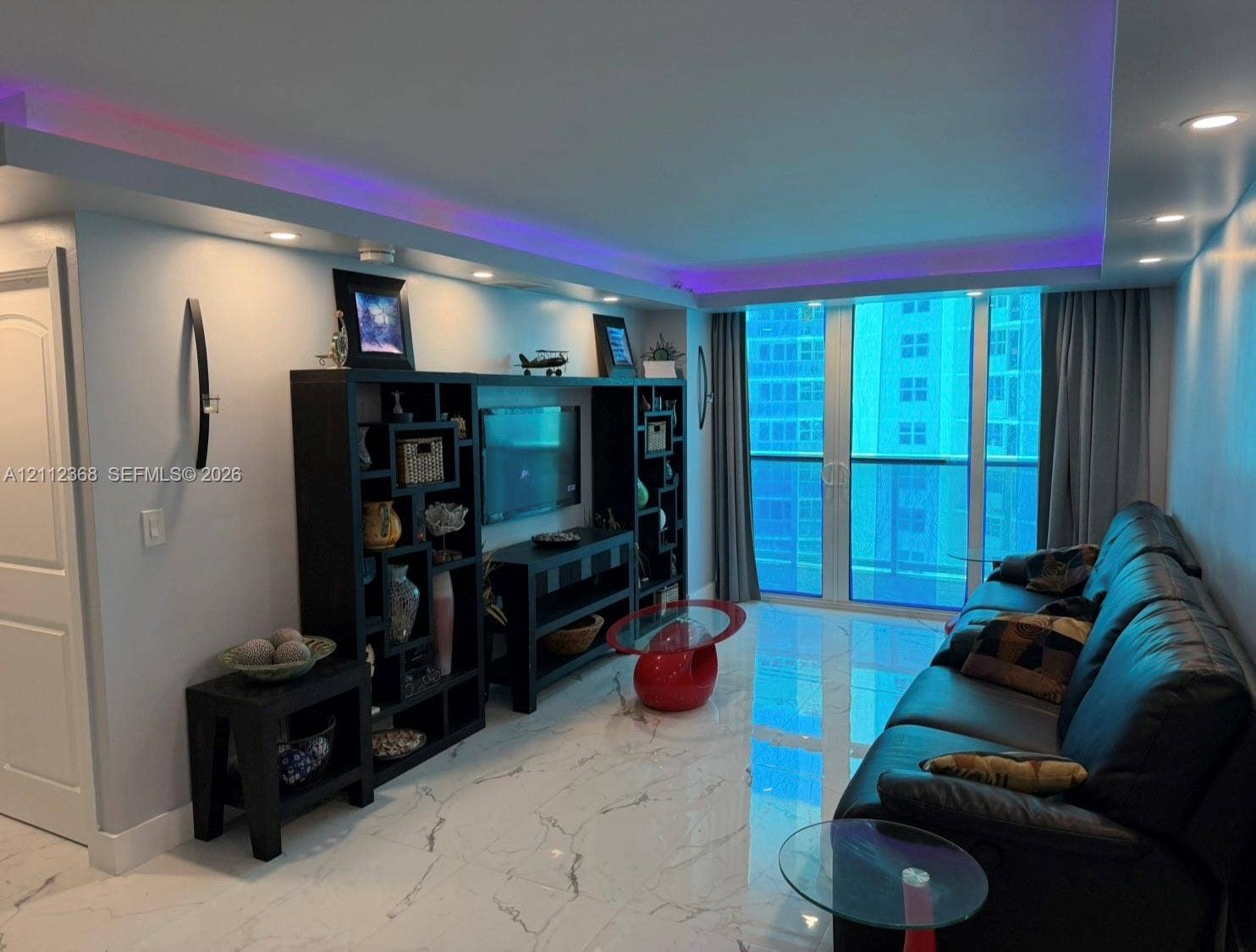 Large-format stone-finish floor tiling, recessed ceiling lighting with integrated colored accent lighting, built-in shelving unit, and floor-to-ceiling glass paneling leading to a balcony
