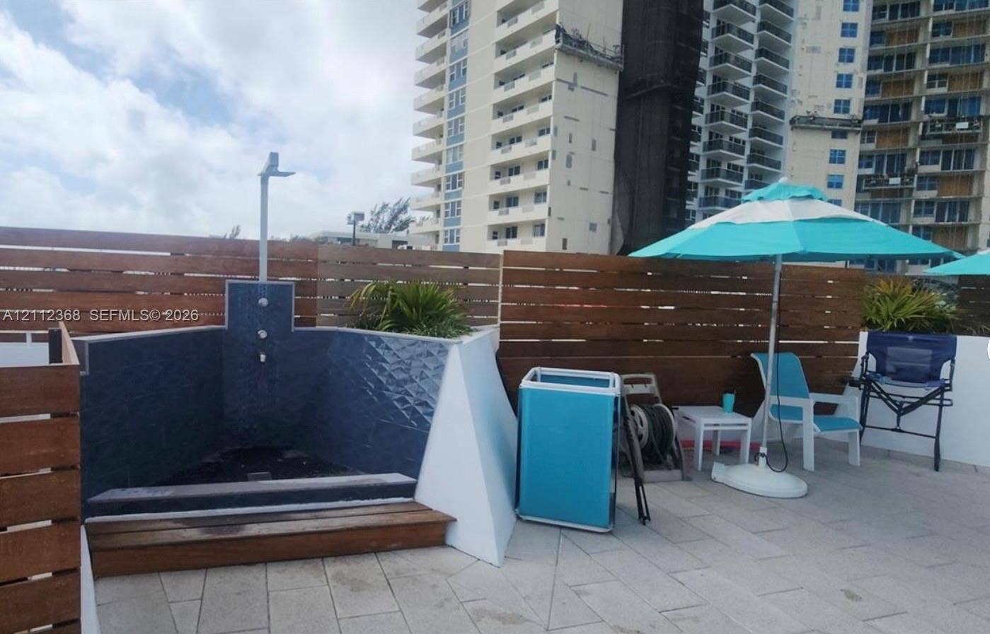 Outdoor shower featuring a rainfall showerhead, navy blue textured tile surround, stone paver flooring, and horizontal wood-slat privacy fencing