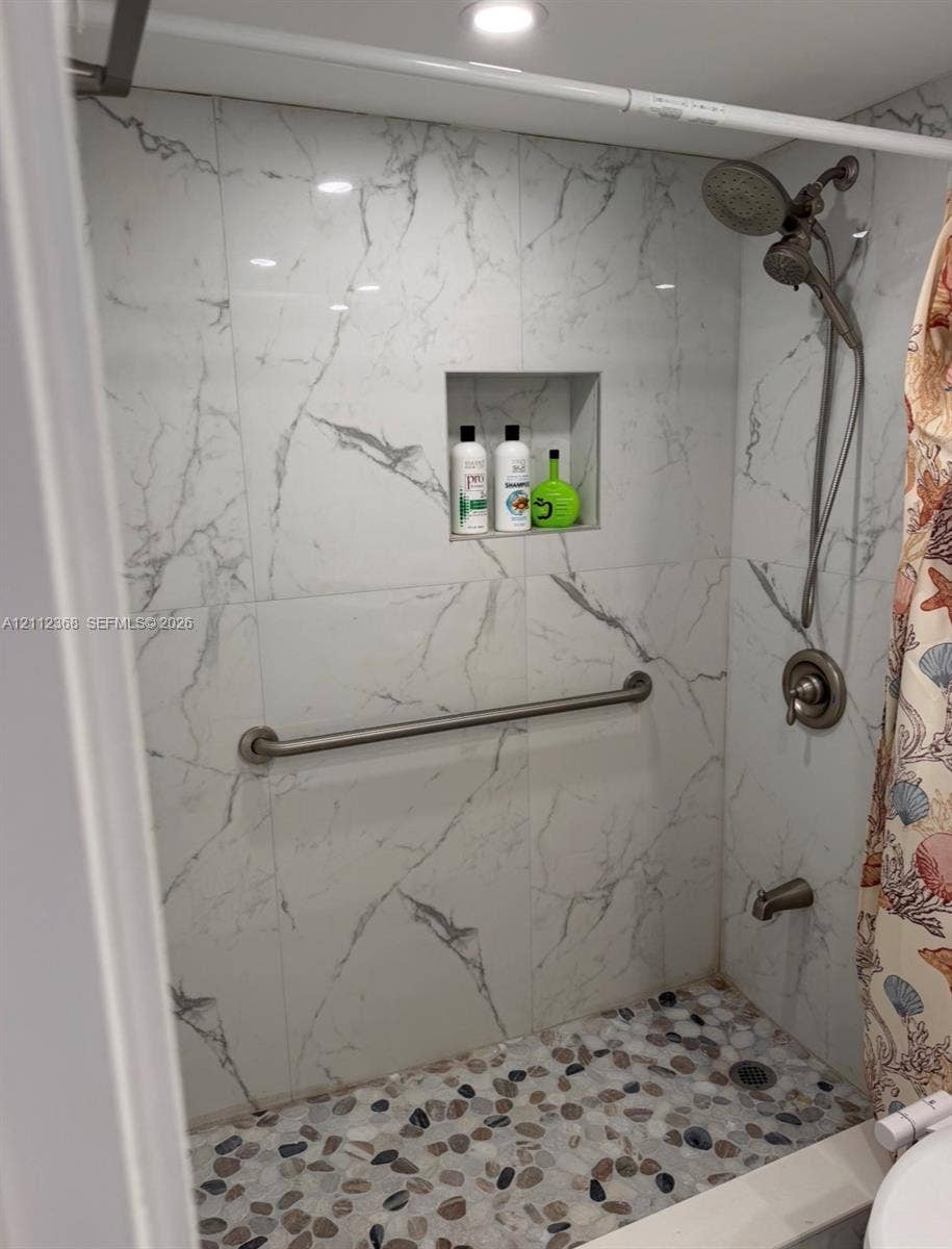 Shower enclosure featuring large-format white stone-finish wall tile, recessed storage niche, pebble-textured flooring, brushed metallic hand-held shower fixture, and wall-mounted grab bar