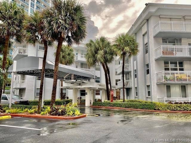Multi-story residential complex featuring covered porte-cochre entry, balconies with railing systems, and landscaped grounds with mature palm trees